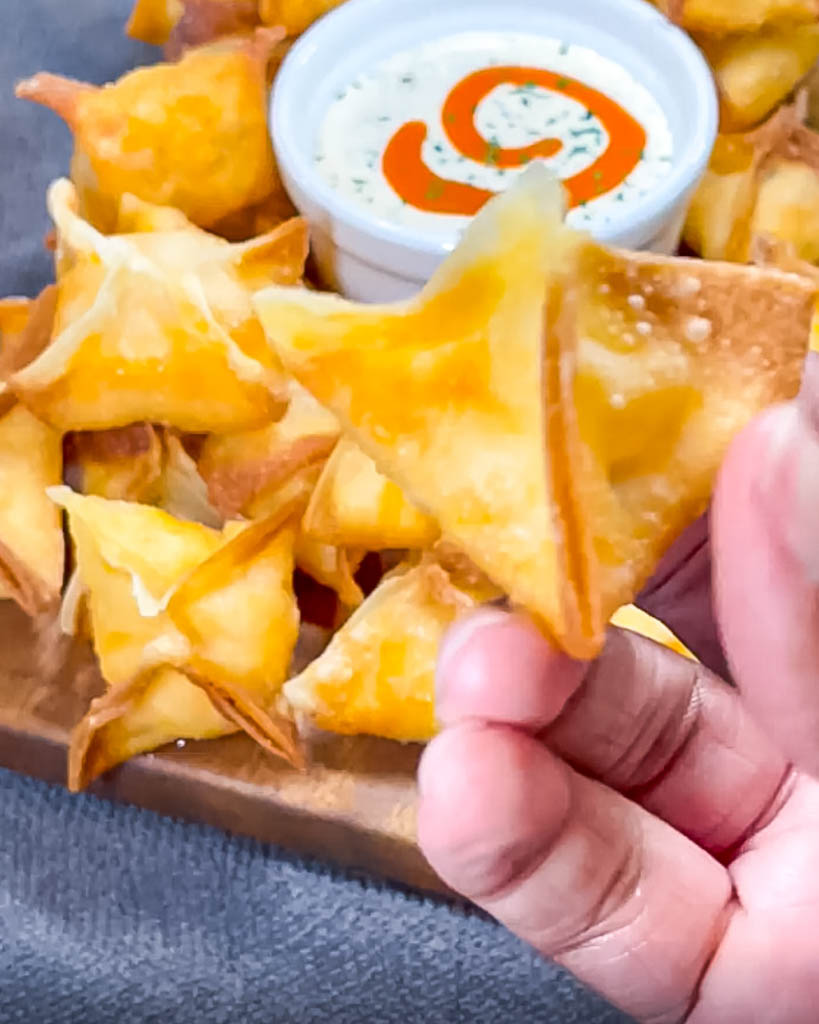 a close up view of a hand holding a buffalo chicken wonton with a platter of more in the background