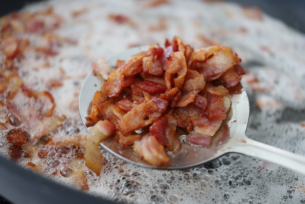 Cut up bacon being fried in a pan. The bacon is being spooned out of the pan for a close up picture.
