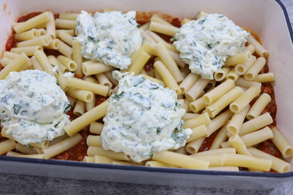 A casserole dish with the meat mixture evenly spread to just cover the bottom of the dish, topped with a layer of cooked penne pasta, and dollops of ricotta mixture.