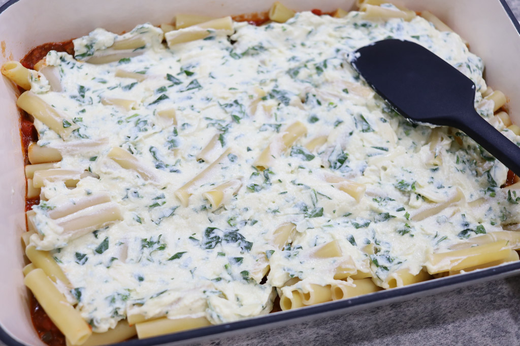 A casserole dish with the meat mixture evenly spread to just cover the bottom of the dish, topped with a layer of cooked penne pasta, and a layer of ricotta mixture. This is being evenly spread with a silicon spoon.