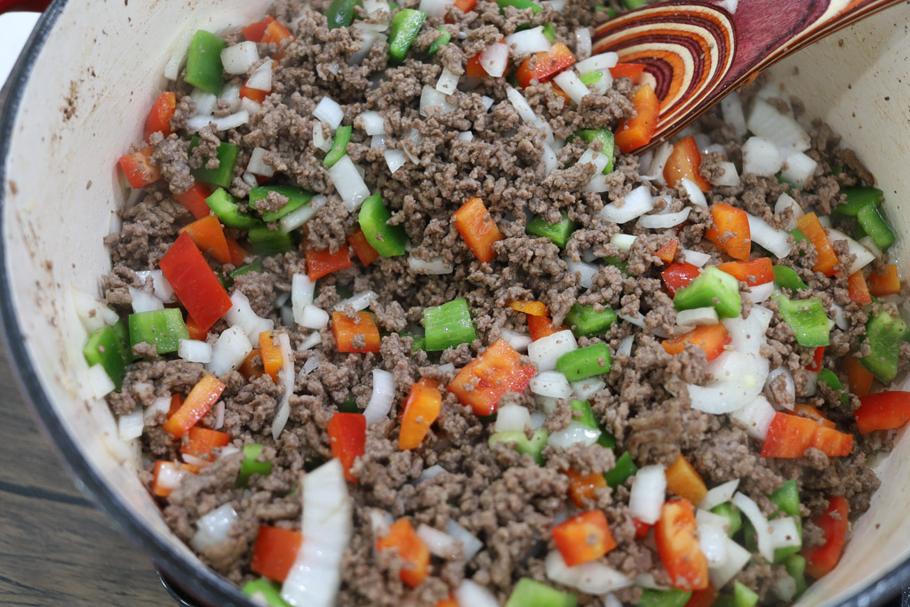 Cooked ground beef mixed with diced onions, green peppers, and red peppers in a dutch oven being stirred with a wooden spoon.