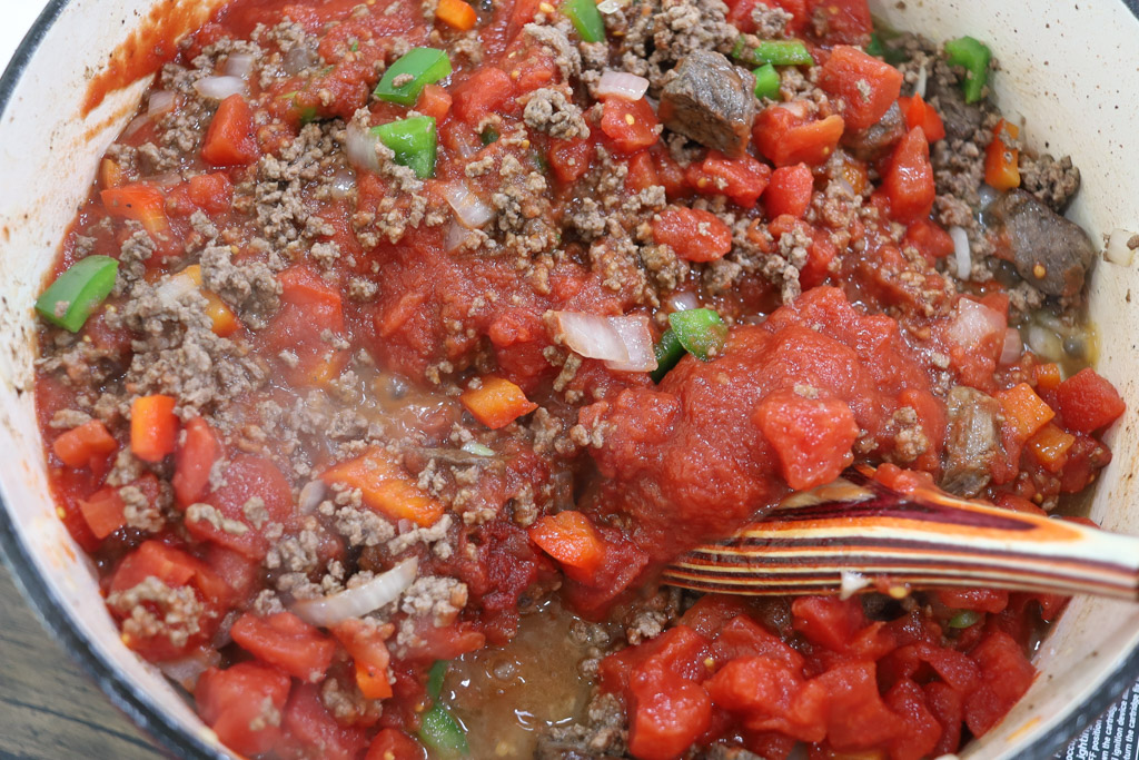Cooked ground beef and diced chuck roast mixed with diced onions, green peppers, and red peppers in a dutch oven being stirred with a wooden spoon. Being mixed in is the diced tomatoes.