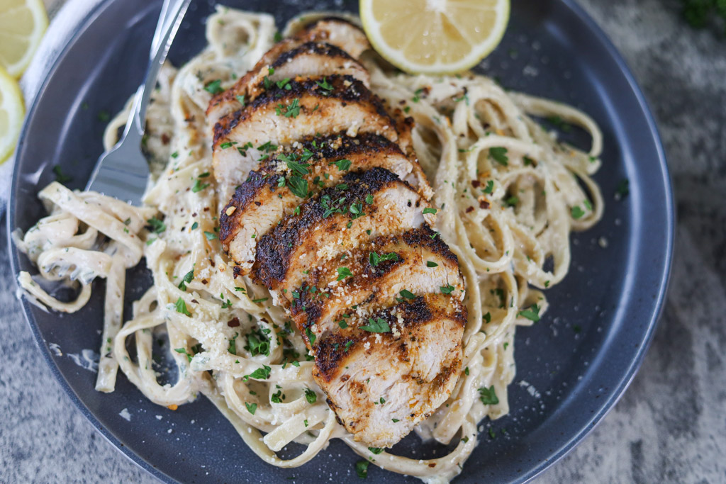 Grilled chicken served on top of fettuccine alfredo noodles on a serving plate with a lemon on the side.