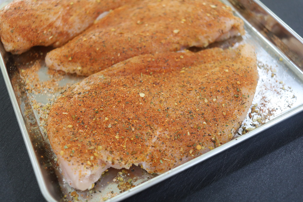 Seasoned, uncooked boneless skinless chicken breasts sitting on a silver baking sheet.