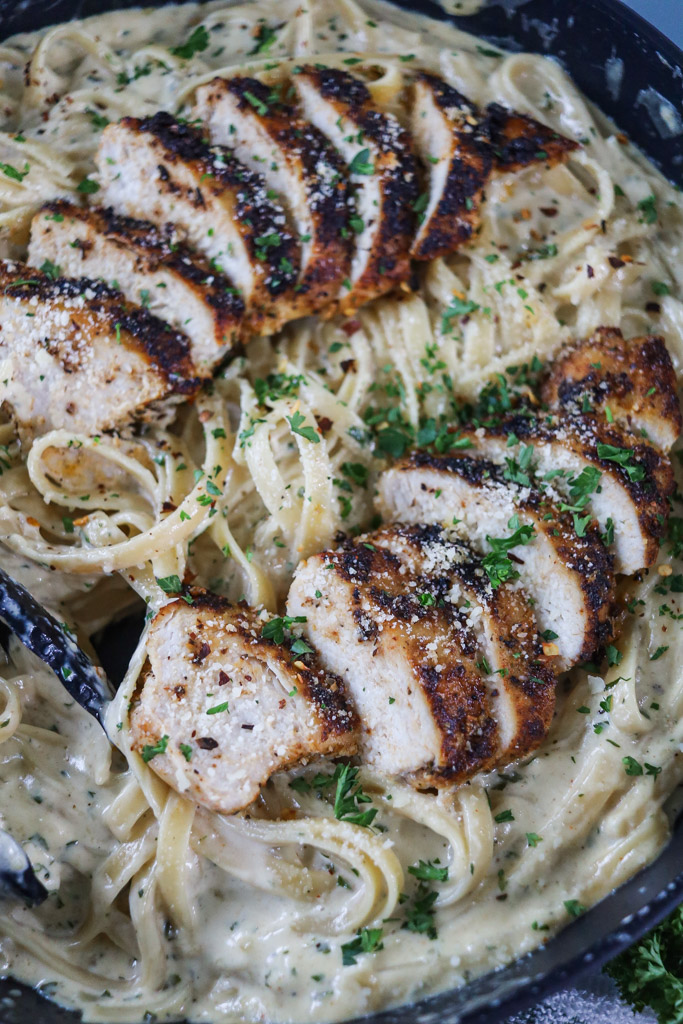 Grilled chicken served on top of fettuccine alfredo noodles on a platter being served with tongs.