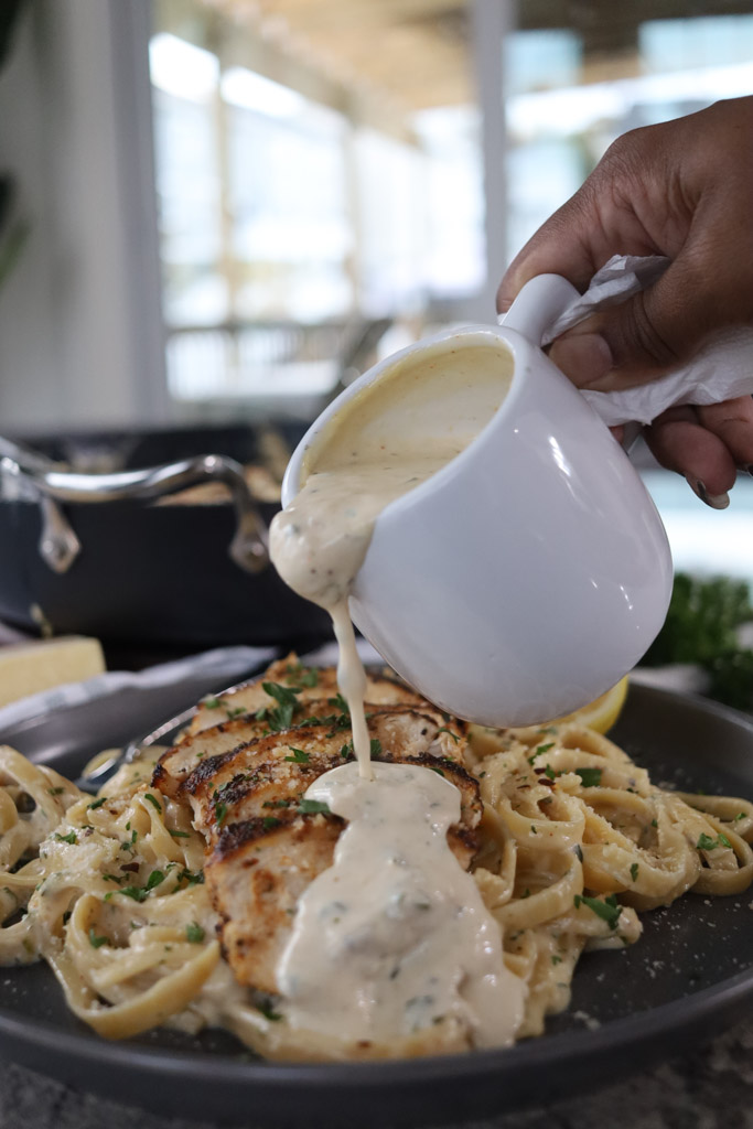Grilled chicken served on top of fettuccine alfredo noodles on a plate being drizzled with more sauce.