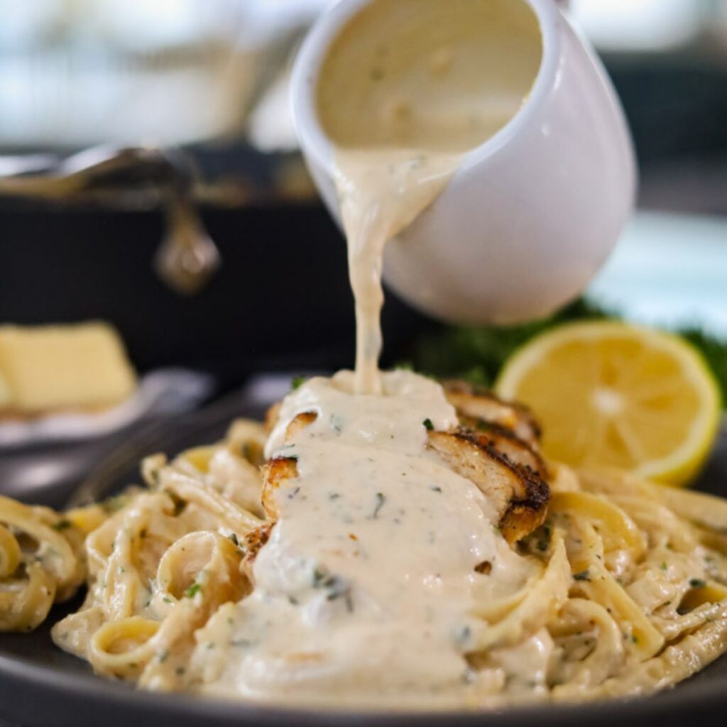 Grilled chicken served on top of fettuccine alfredo noodles on a plate being drizzled with more sauce.