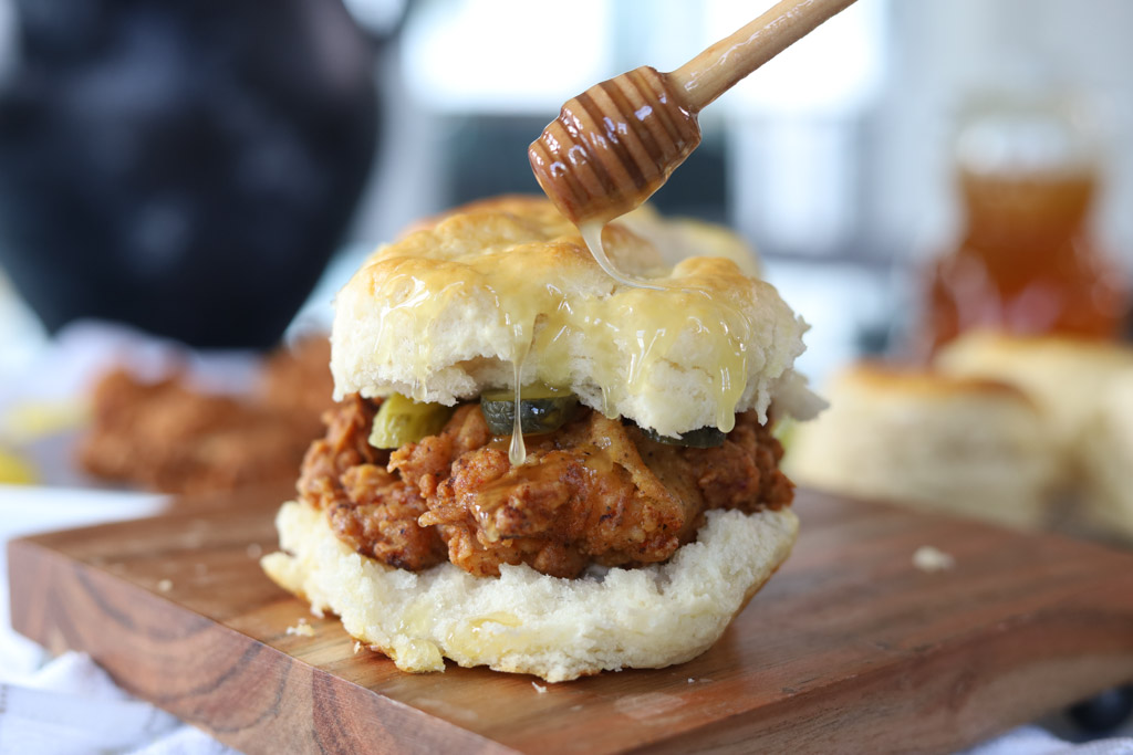Honey butter chicken biscuit being shown on a wooden cutting board with a honeycomb utensil drizzling sauce over the top.
