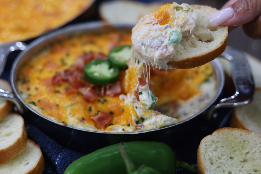 Jalapeno popper dip in a serving dish with crostinis and jalapenos on the side. Showing dip in detail with close up of someone dipping into the appetizer.