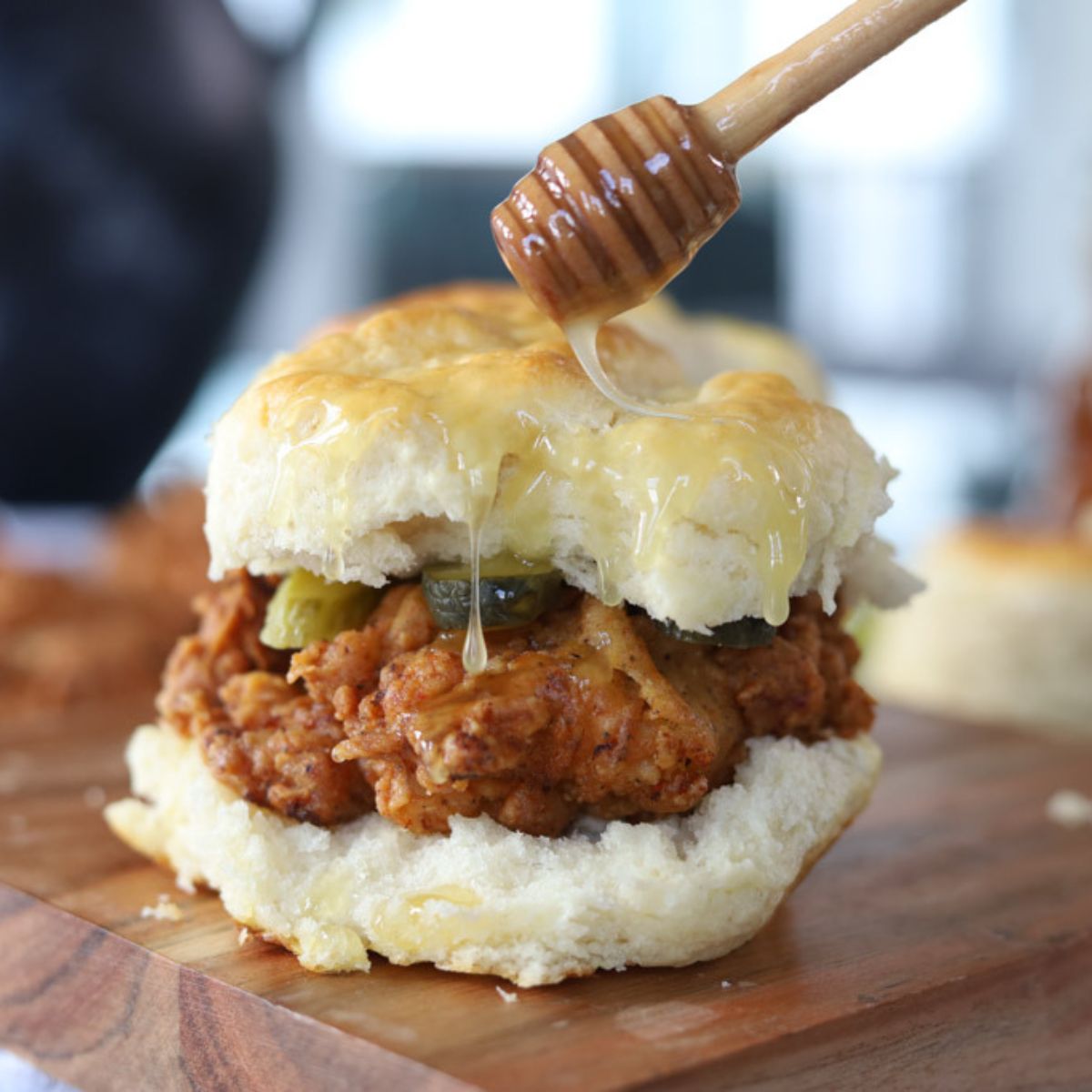 Honey butter chicken biscuit being shown on a wooden cutting board with a honeycomb utensil drizzling sauce over the top.