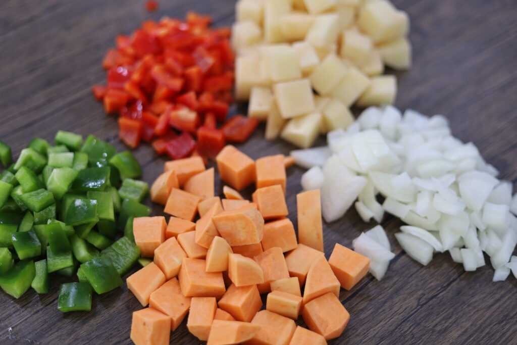 Uncooked, diced ingredients needed for the breakfast skillet on a wooden cutting board including onions, sweet potatoes, potatoes, red pepper, and green pepper.