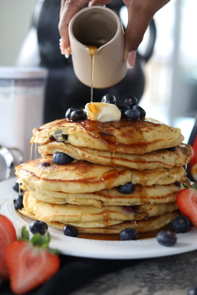 fluffy blueberry pancakes stacked high topped with blueberries, butter, and syrup alongside strawberries.