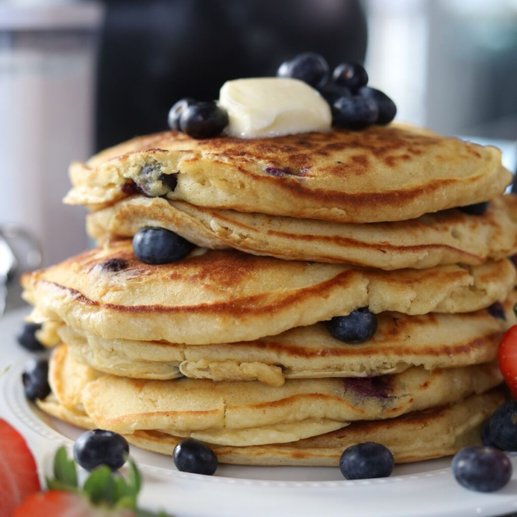 fluffy blueberry pancakes stacked high topped with blueberries and butter alongside strawberries.
